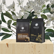 Two packets of Kruti Coffee, one labeled as 'South Indian Filter Coffee' and the other as 'Premium Blend', against a background with white flowers and green leaves.